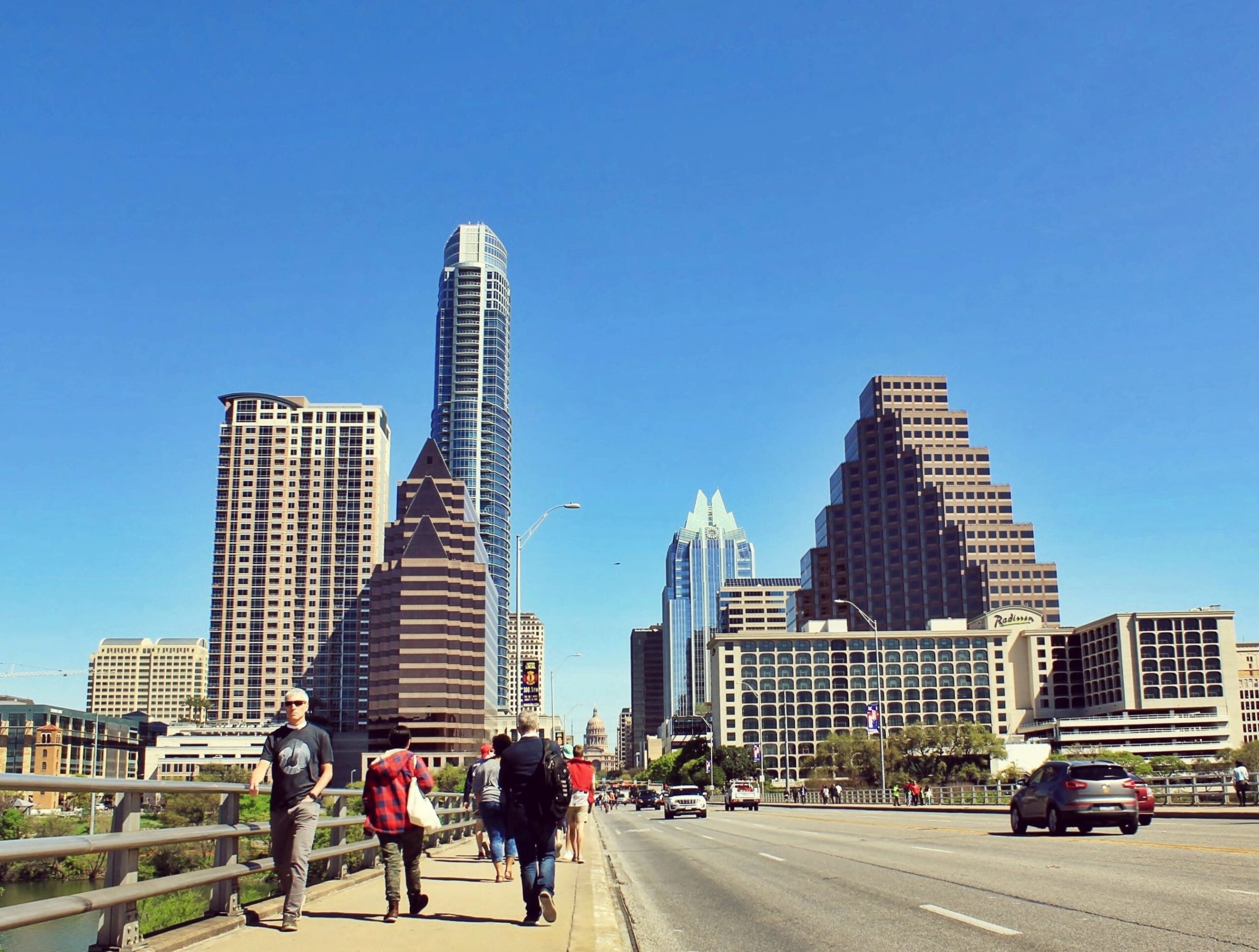 Austin skyline against a blue sky with three adults walking across a bridge and cars passing on the roadway
