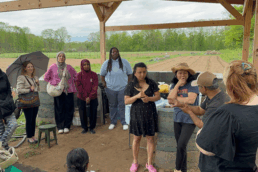 Standing outside in a field rimmed with trees, partly sheltered by an open wooden structure, a group of five PLACES Fellows, one carrying an umbrella, stand in a semicircle around three women from the Asha Laaya Farm. The women are communicating via sign language.. A red-headed person in a black shirt stands in the foreground with their back to the camera.